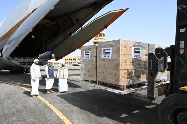 Three men standing on an airfield next to boxes of cargo being unloaded from an airplane.