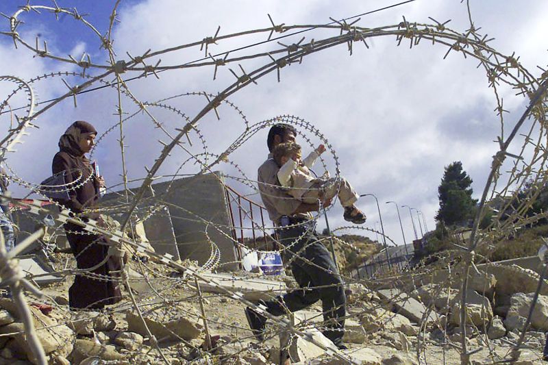 رجل فلسطيني يحمل طفله لعبور حاجز طريق في مدينة الخليل بالضفة الغربية في عام 2003. ويملأ الركام الأرض التي يسير عبرها وتلتف الأسلاك الشائكة أمامه.  وتتبعهم امرأة ترتدي غطاء الرأس.