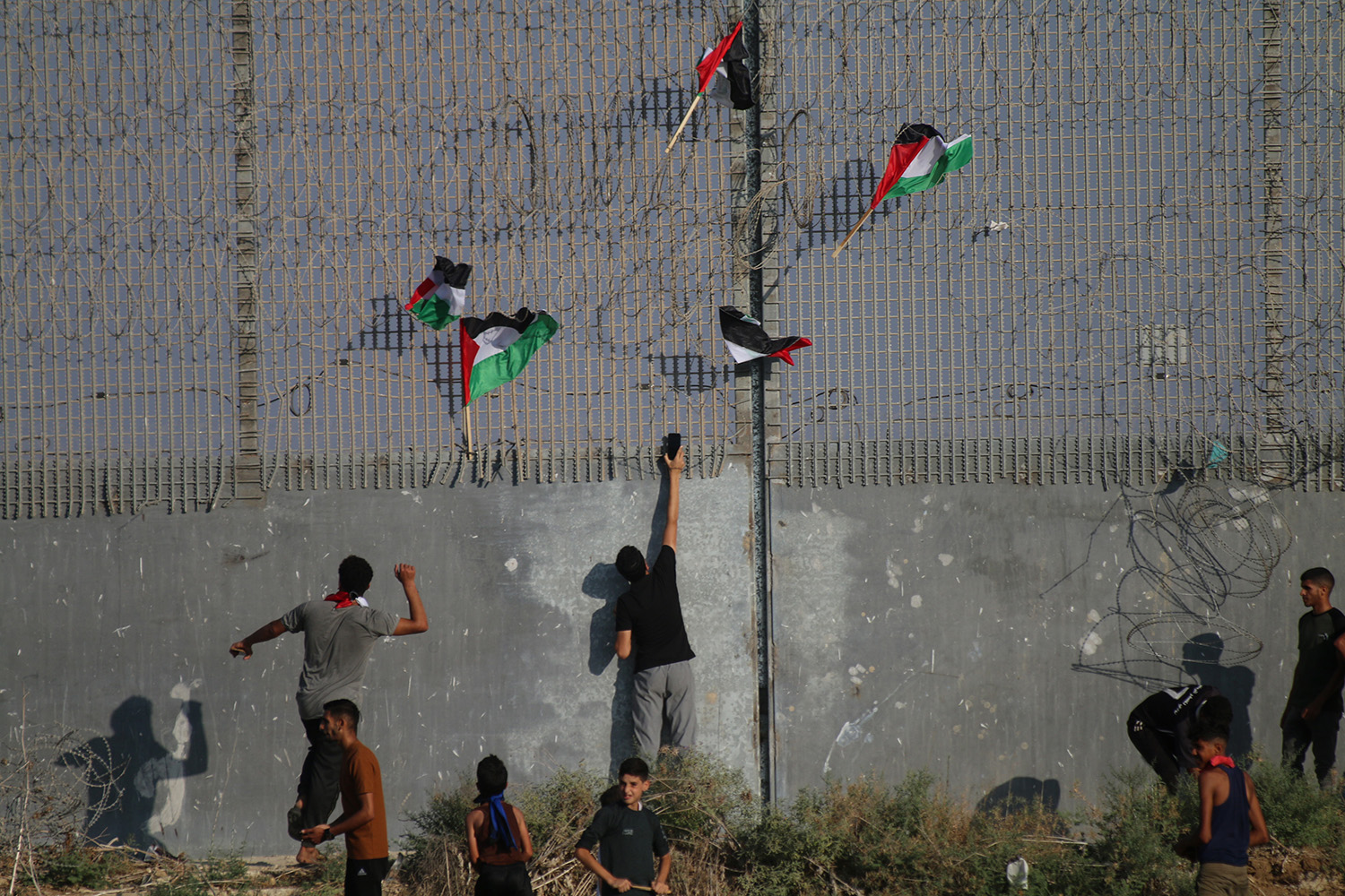 متظاهرون فلسطينيون يتجمعون بالقرب من السياج الحدودي بين قطاع غزة وإسرائيل ويتسلقونه، وأعلام إسرائيل عالقة في الأسلاك الشائكة للسياج.