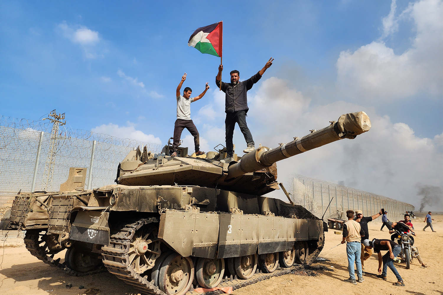 أعضاء في الجناح العسكري لحركة حماس يرفعون العلم الفلسطيني فوق دبابة إسرائيلية.  ويمتد خلفهم سياج حدودي على مسافة.